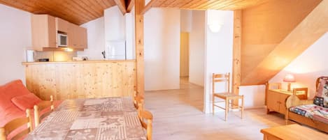 A cozy room with a wooden ceiling, a dining table with chairs, a small kitchen area, and a separate seating area with a lamp.