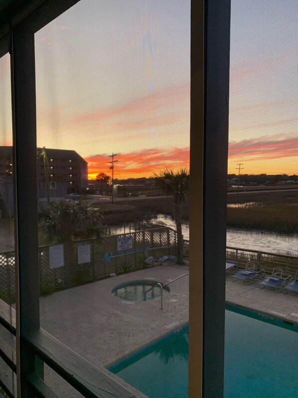 A view of a swimming pool with a fence and lounge chairs, a building in the background, and a sunset sky. - Garden City Beach- Great Location! 2 minute walk to beach and pier! Sleeps 6.  (Murrells Inlet)