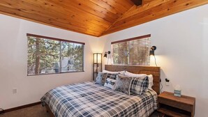 Un dormitorio con techo de madera, una cama con colcha escocesa, dos lámparas de noche y una mesita de noche con una caja de pañuelos.