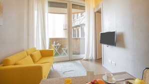 A room with a yellow sofa, a flat-screen TV mounted on the wall, a small table with a cup and a plate, and a view of a balcony with a table and chairs outside. - Sweett | Torre Velasca I (Milano)