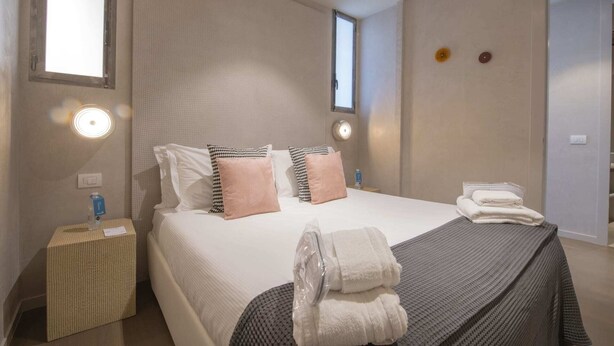 A neatly arranged hotel room with a large bed, two bedside tables, a small window, and a door. - Sweett | Torre Velasca VI (Milano)