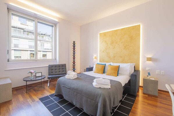 A modern hotel room with a large bed, a bedside table, a chair, a small table with a vase, and a window with a view of buildings. - Sweett | Torre Velasca VII (Milano)