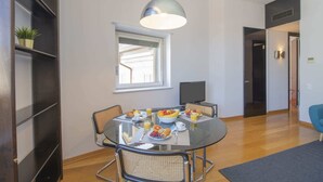 Un comedor con una mesa redonda, sillas e iluminación colgante. Se aprecia una ventana con persianas, un televisor y una puerta que da a otra habitación.