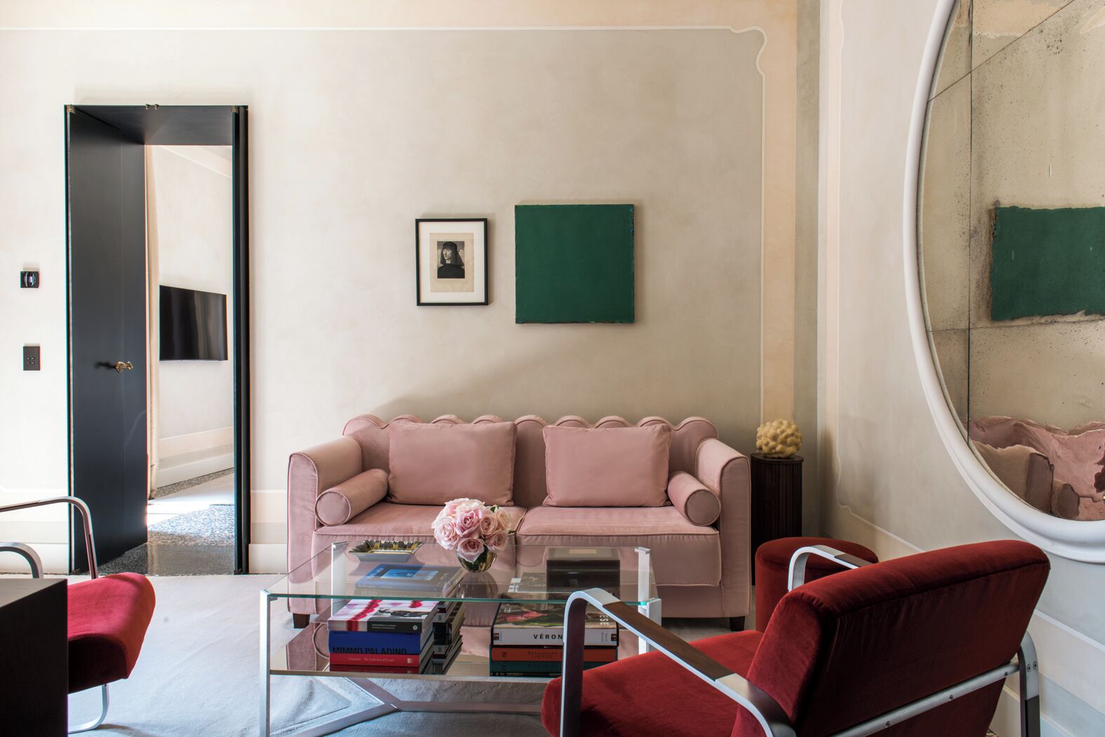 Un salon moderne avec un canapé rose, une table basse en verre sur laquelle sont posés des livres, et des fauteuils rouges.