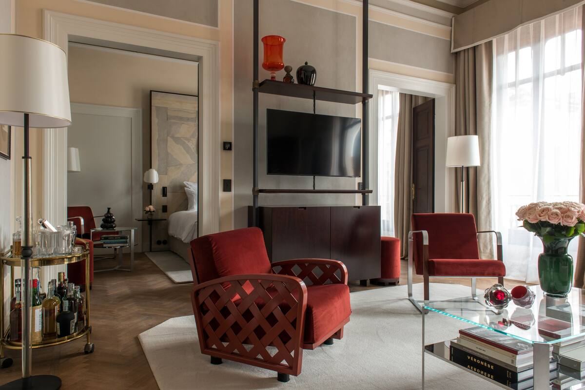 A modern hotel room with a large flat-screen TV, red upholstered chairs, a glass coffee table, and a bookshelf.