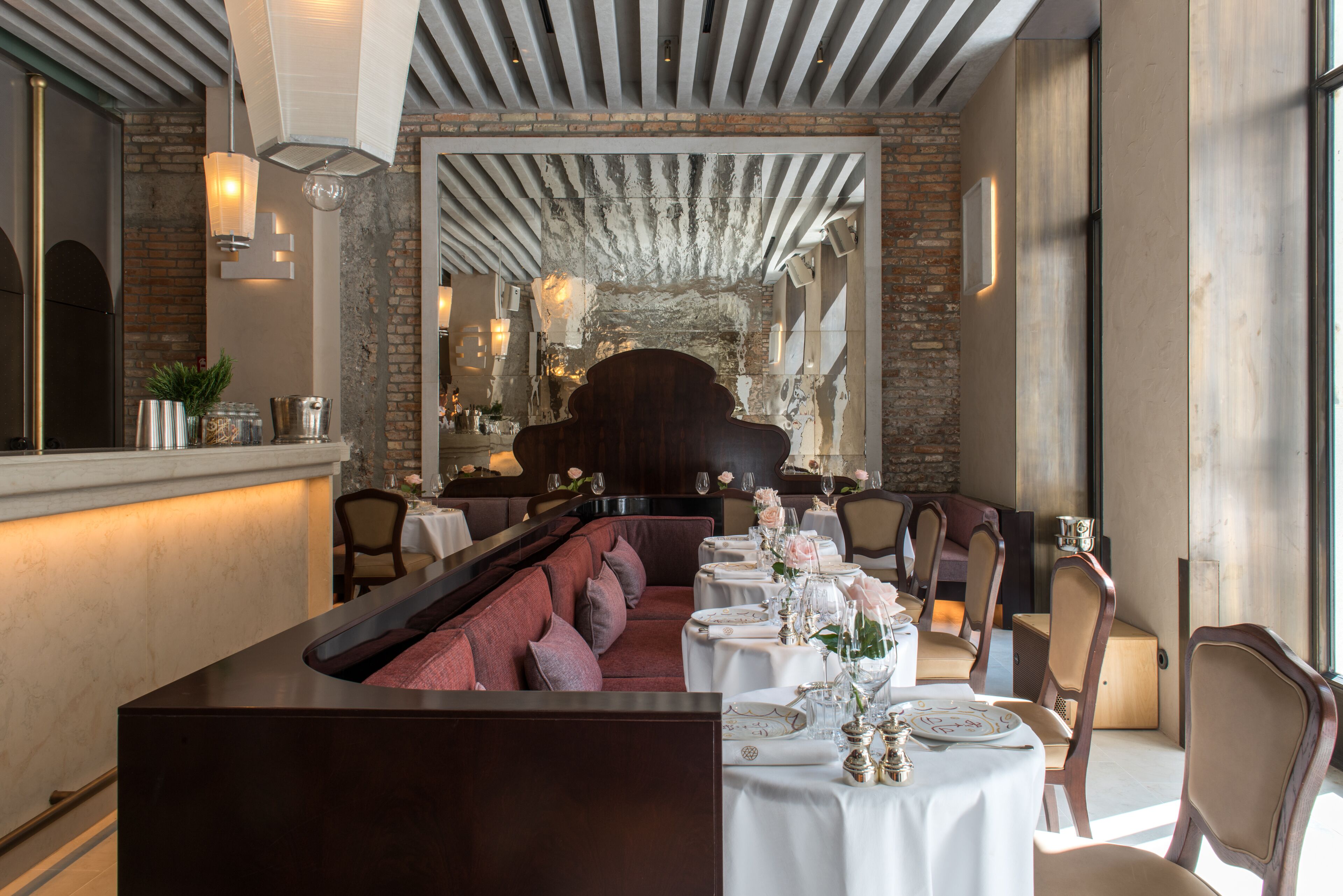 Um restaurante com uma mesa de jantar comprida posta para uma refeição, um sofá vermelho e uma parede de tijolos ao fundo.