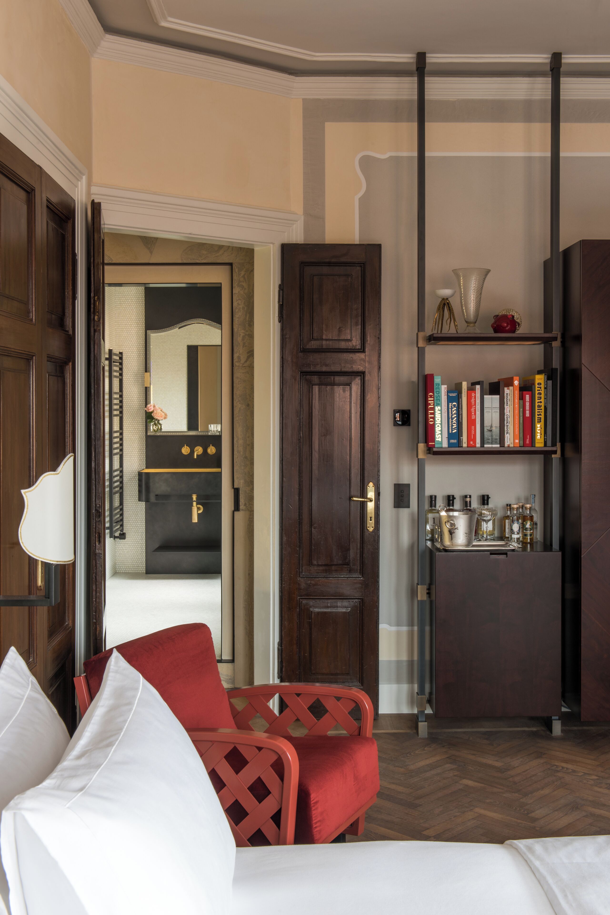 Habitación de hotel con puerta de madera, una estantería llena de libros, una pequeña zona de cocina con hervidor y una silla roja.