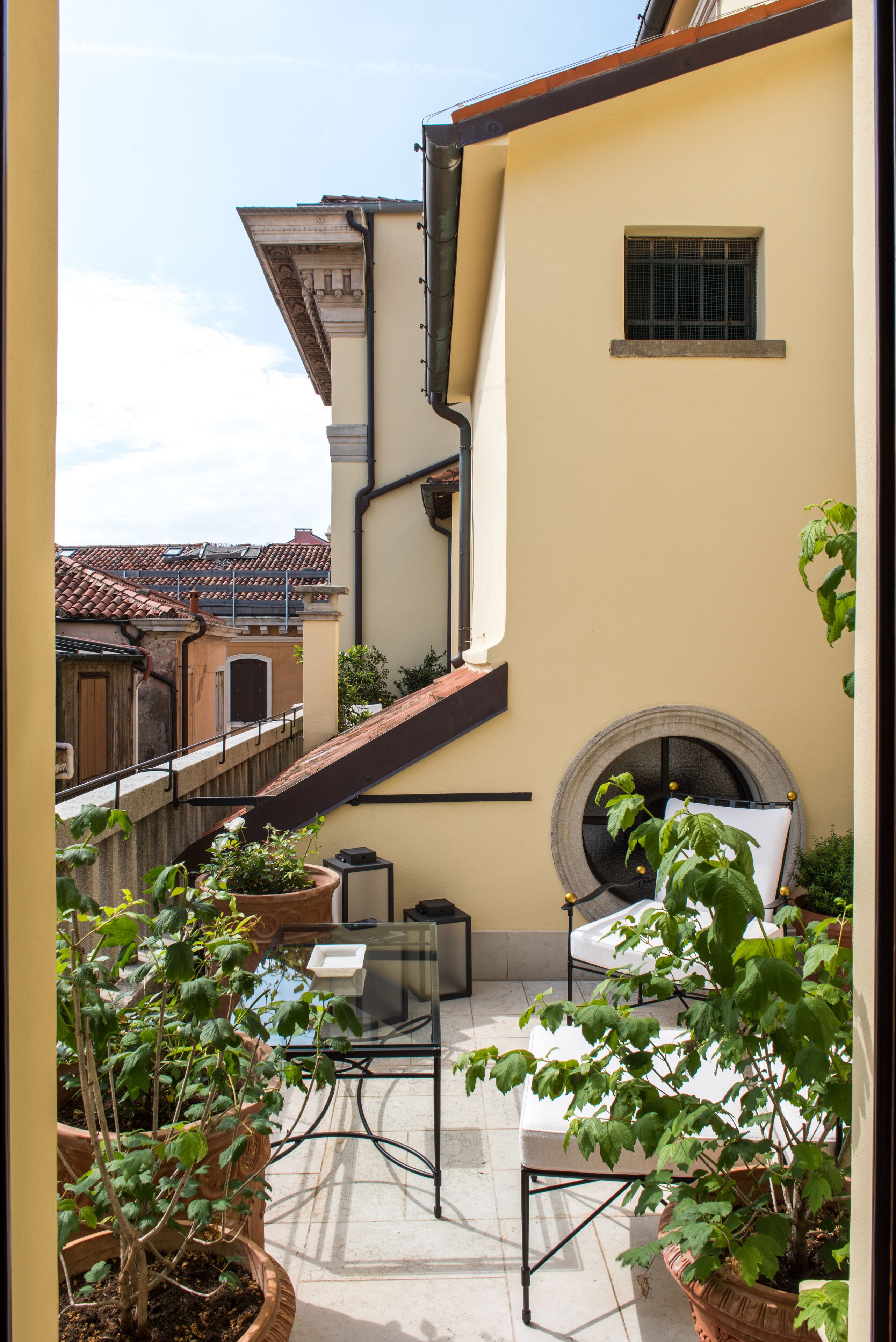 A balcony with a table, chairs, potted plants, and a view of neighboring buildings.