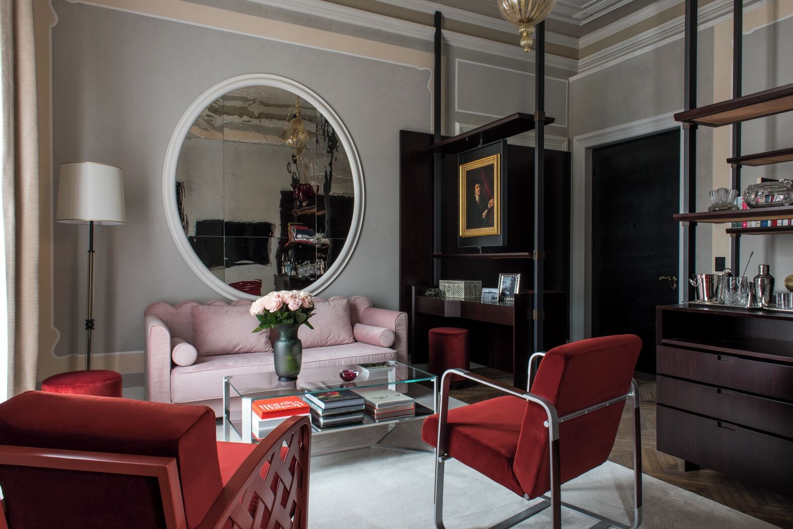 Una sala de estar moderna con un espejo redondo, un sofá rosa, sillas rojas, una mesa de centro de vidrio y una estantería de madera oscura.
