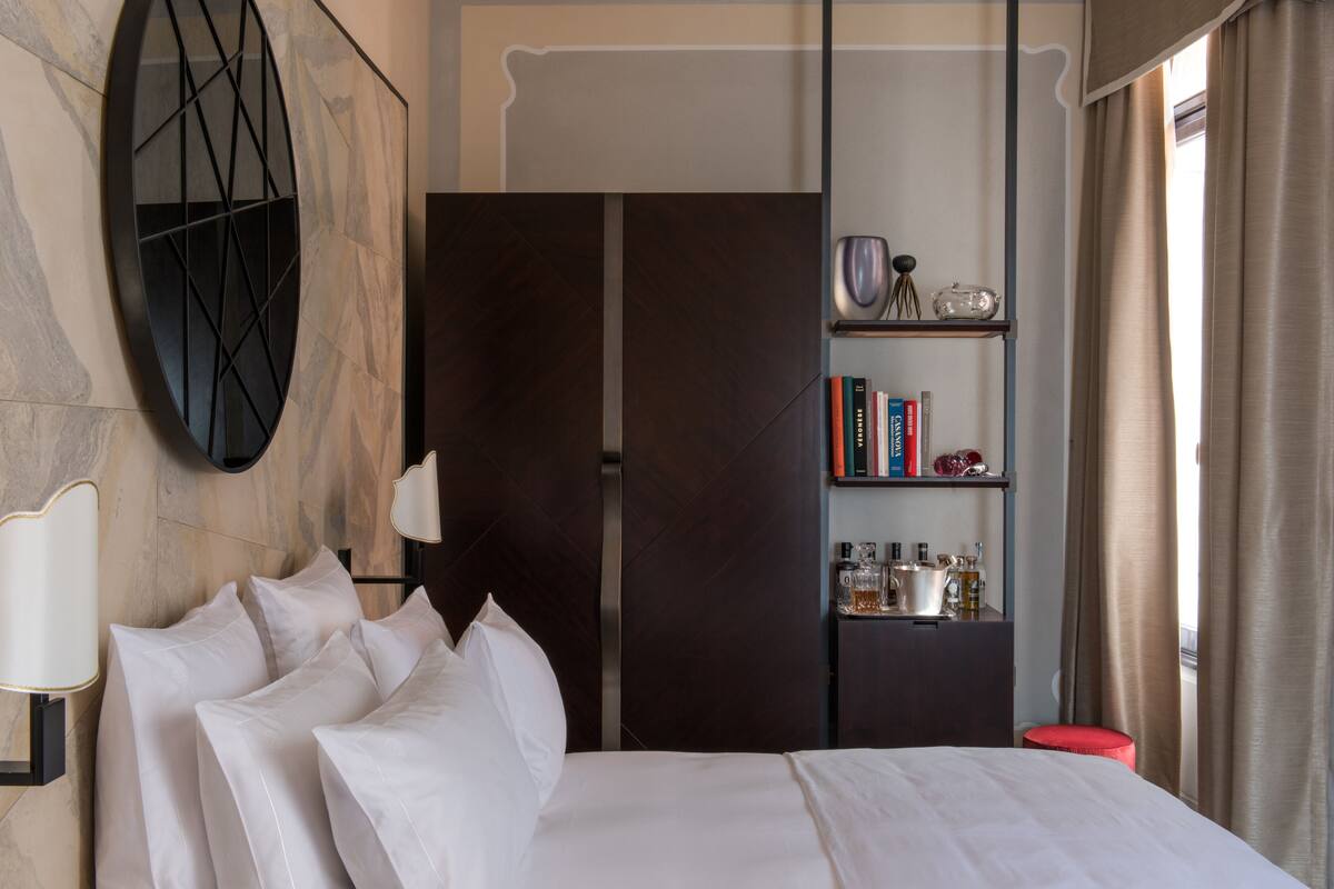 A hotel room with a bed, a bookshelf, a mirror, and a red ottoman.