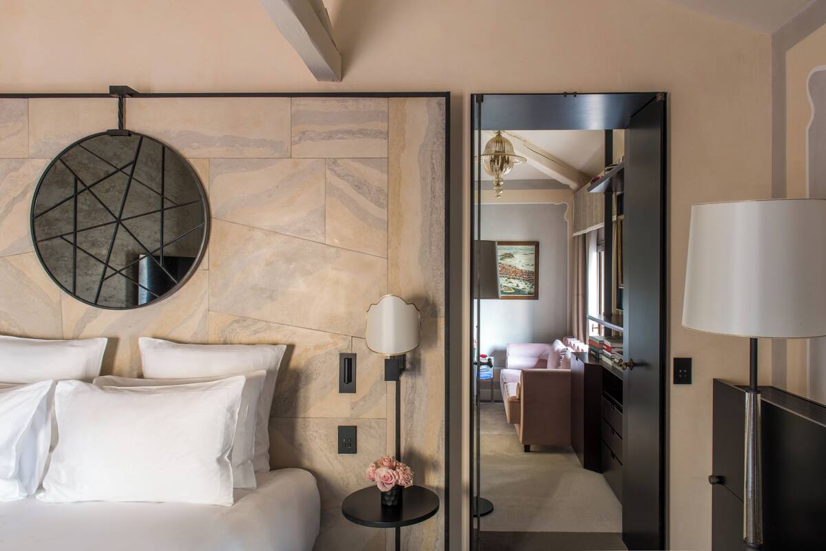 A hotel room with a bed, a round mirror, a lamp, and a view into the adjacent room.