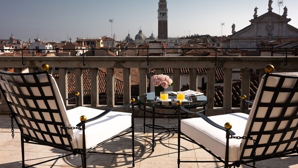 A terrace with two white cushioned chairs, a glass table with a vase of flowers, and a view of historic buildings including a tall tower. - Nolinski Venezia - Evok Collection (Venice)