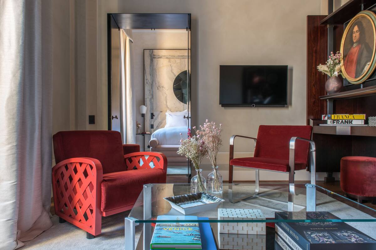 A modern hotel room with a glass coffee table, red upholstered chairs, a bookshelf, and a flat-screen TV.