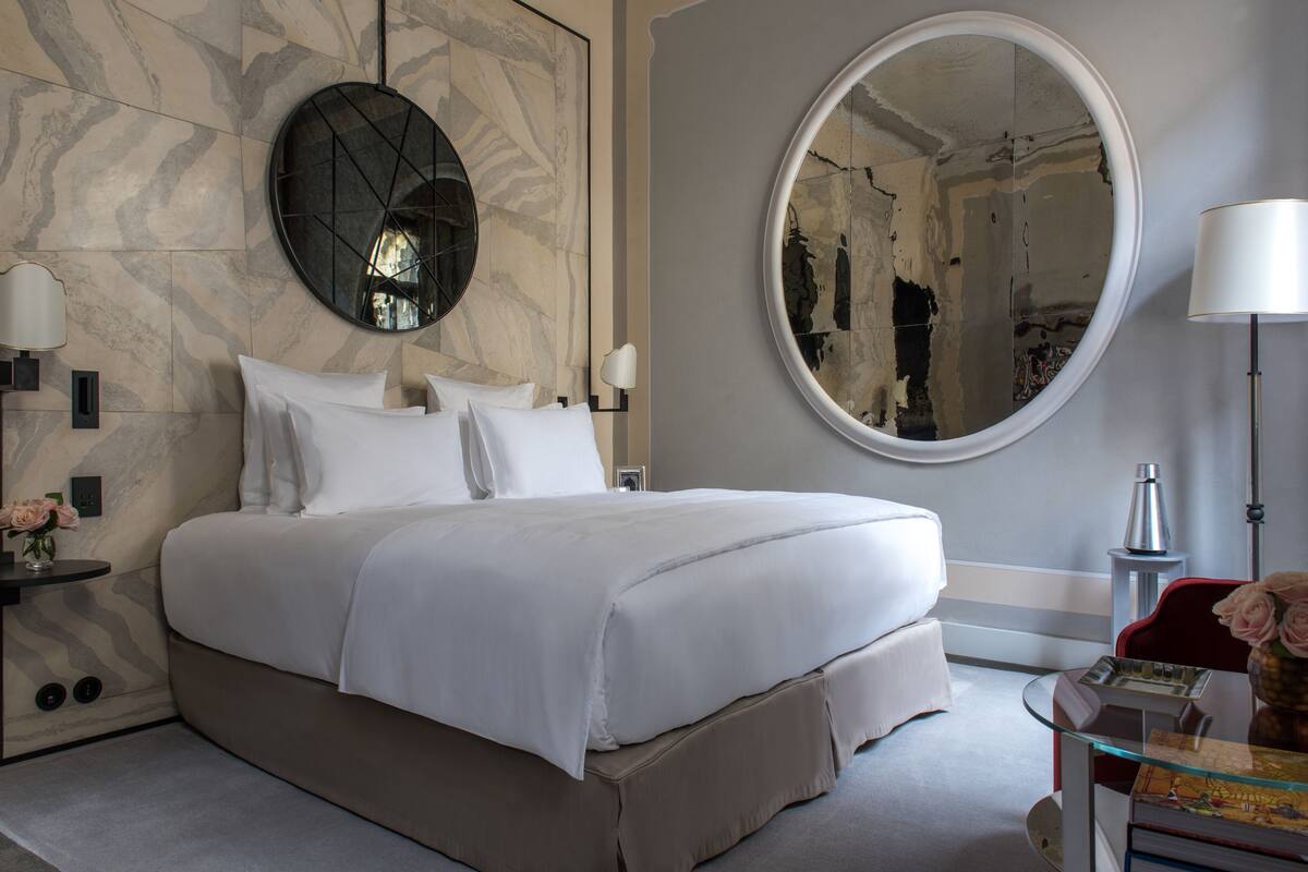 A modern bedroom with a large bed, two wall-mounted lamps, a round mirror, and a glass side table with a vase of flowers.