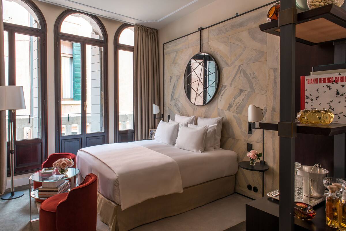 A hotel room with a large bed, a red chair, a small table with books and flowers, and a round mirror on the wall.