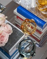 Un objeto de vidrio decorativo con superficie reflectante, sobre una superficie con libros y un pequeño jarrón de rosas rosadas.