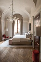 Um quarto espaçoso com uma cama grande, um candelabro, uma poltrona vermelha e piso de madeira em padrão herringbone.