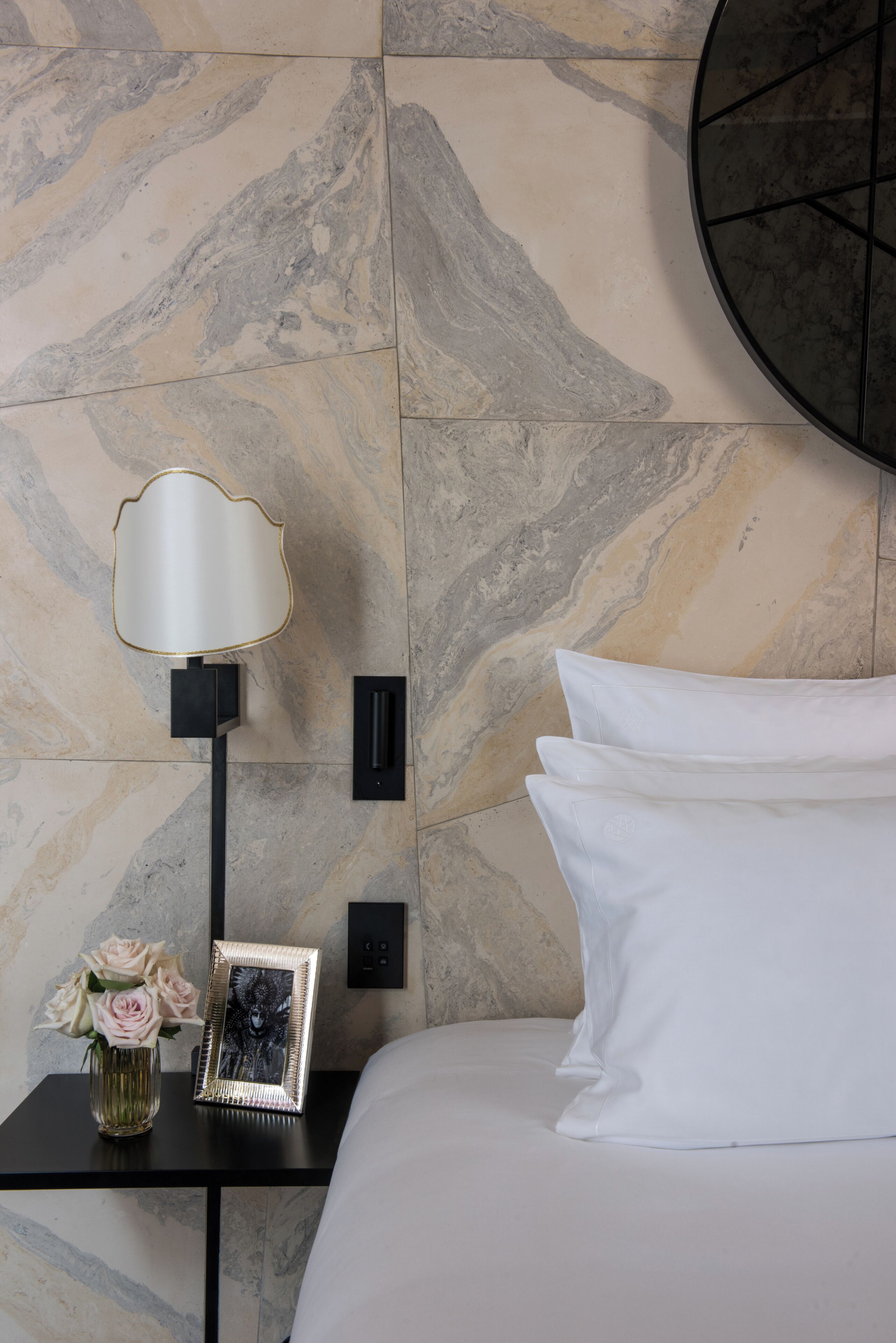 A neatly made bed with white pillows and a black nightstand beside it. On the nightstand, there is a lamp with a white shade, a framed picture, and a vase with flowers. The wall behind has a marble-like pattern.