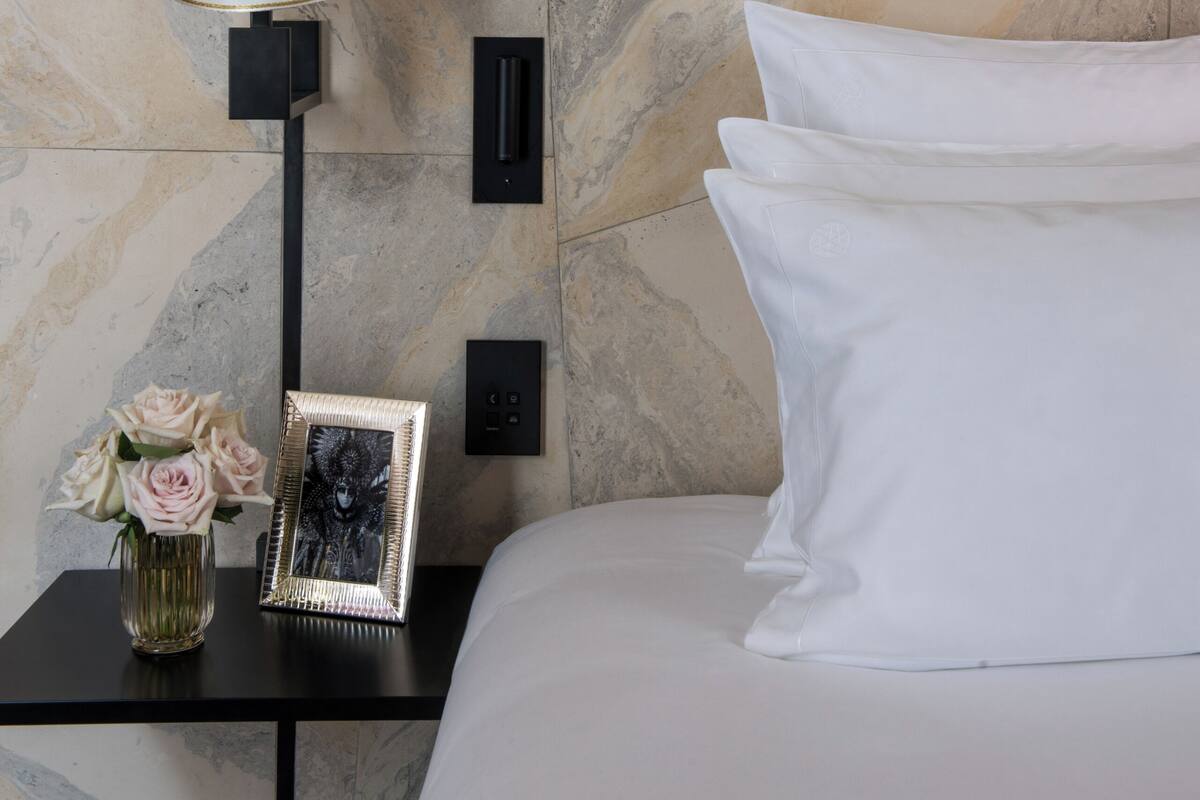 A neatly made bed with white pillows and a black nightstand beside it. On the nightstand, there is a lamp with a white shade, a framed picture, and a vase with flowers. The wall behind has a marble-like pattern.