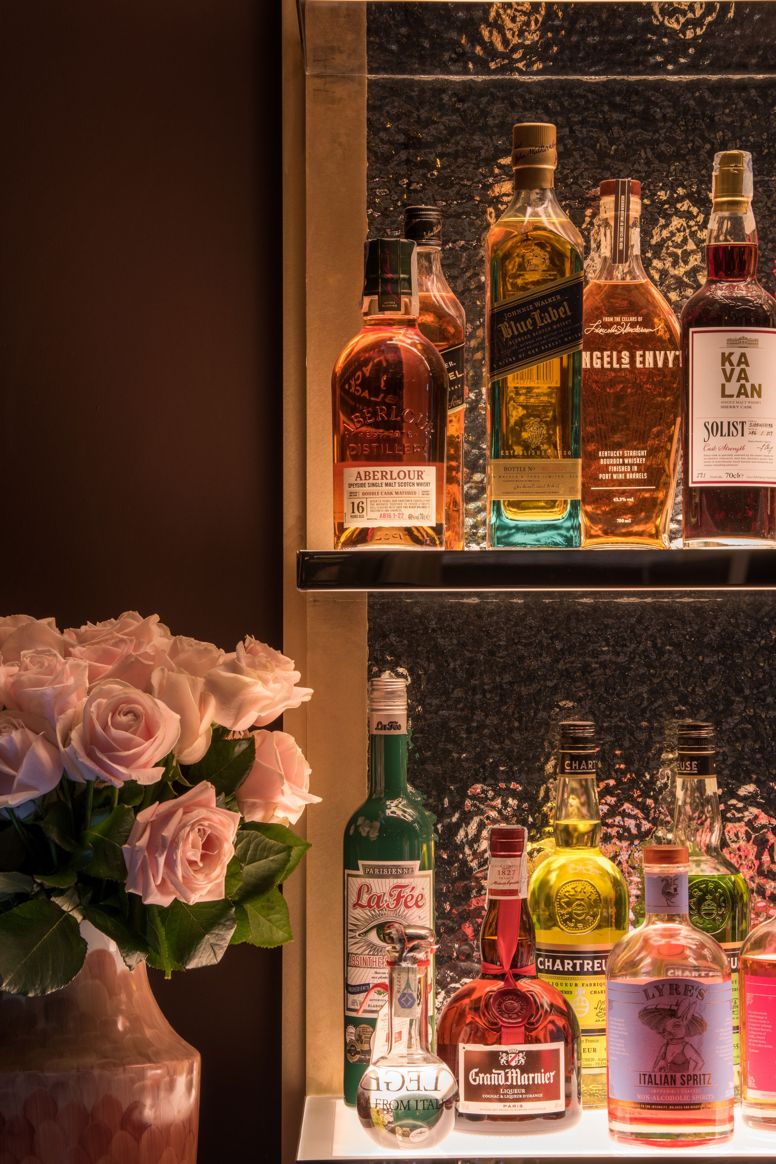 A variety of liquor bottles displayed on shelves, with a vase of pink roses in the foreground.