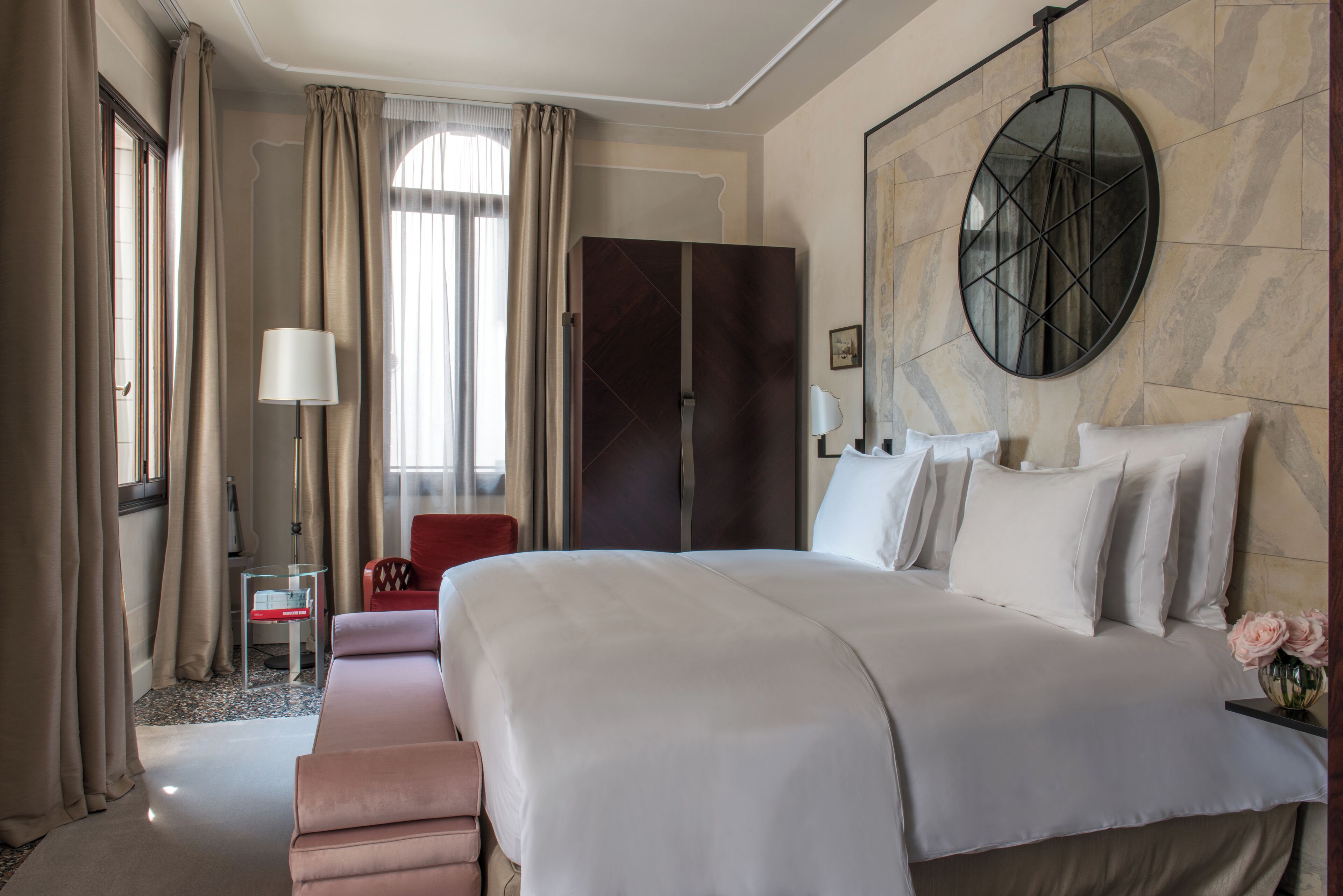 Habitación de hotel con una cama grande, una silla roja, una mesita y un espejo grande en la pared.