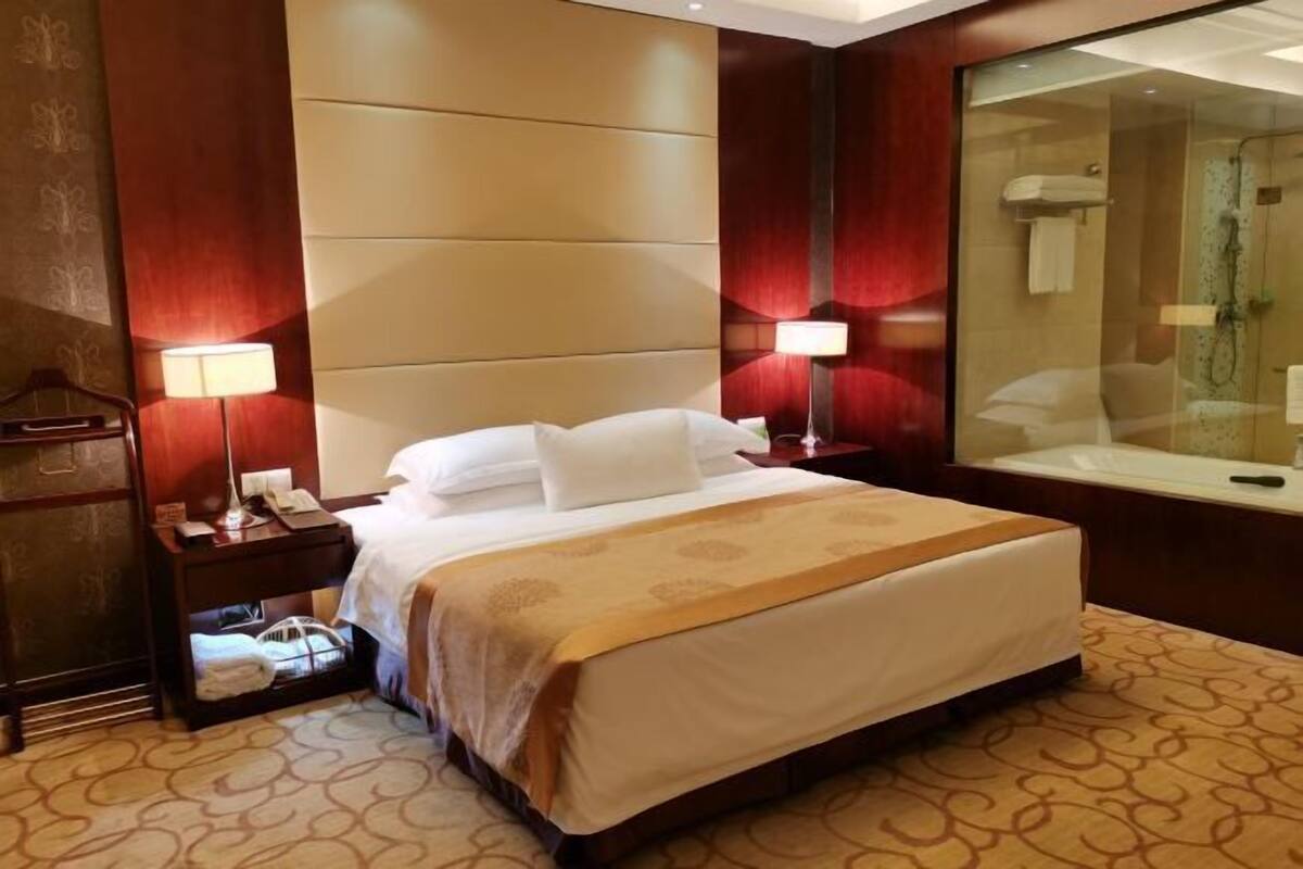 A hotel room with a large bed, two bedside lamps, a nightstand with a phone, a mirror, and a shower area.
