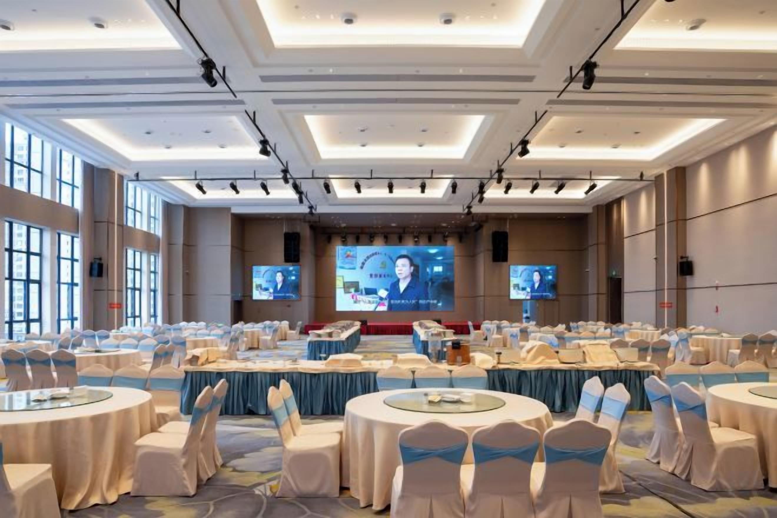 A large conference room with multiple tables, chairs, and a stage with a large screen.