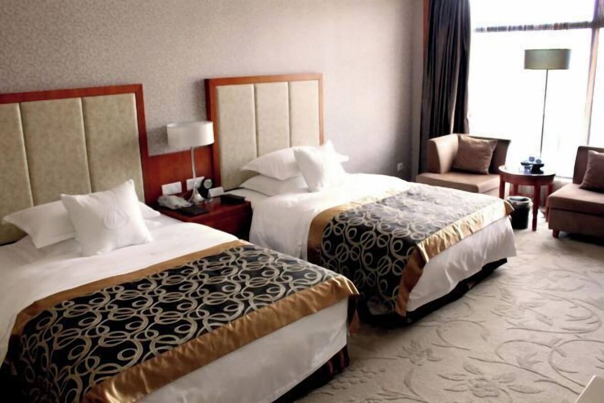A hotel room with two beds, a nightstand, a lamp, a chair, and a view of the outdoors.