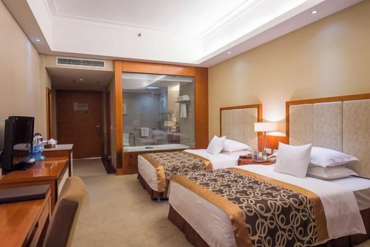 A hotel room with two beds, a desk with a computer, and a bathroom visible through a glass door.