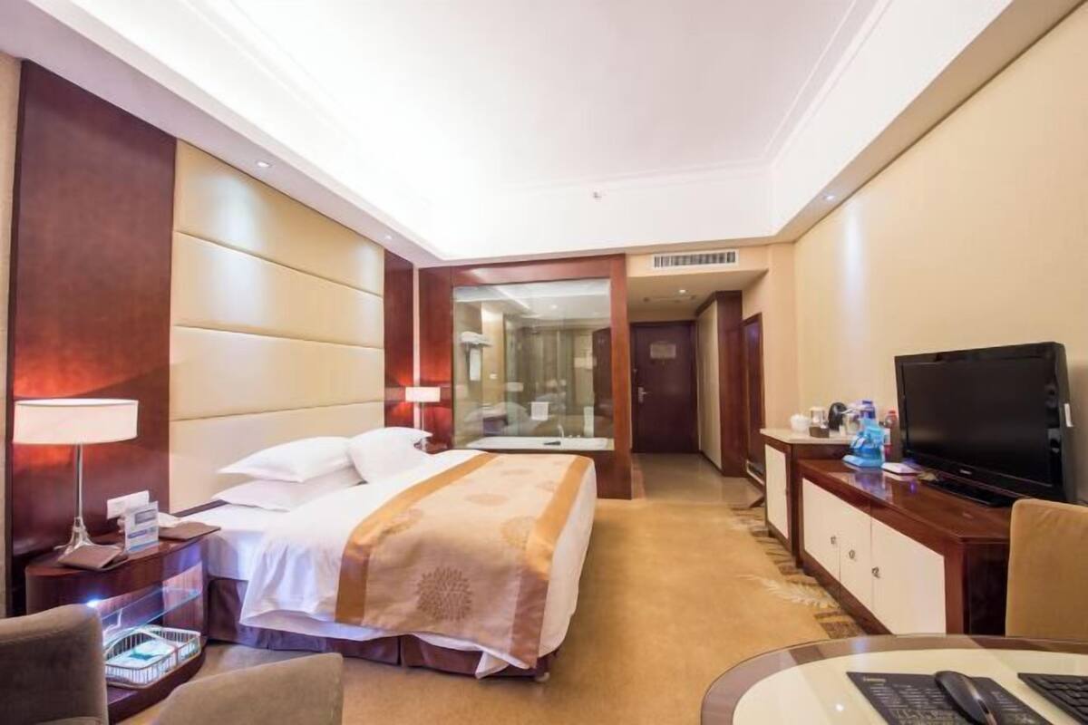A hotel room with a bed, bedside table, lamp, TV, and a desk with a chair.