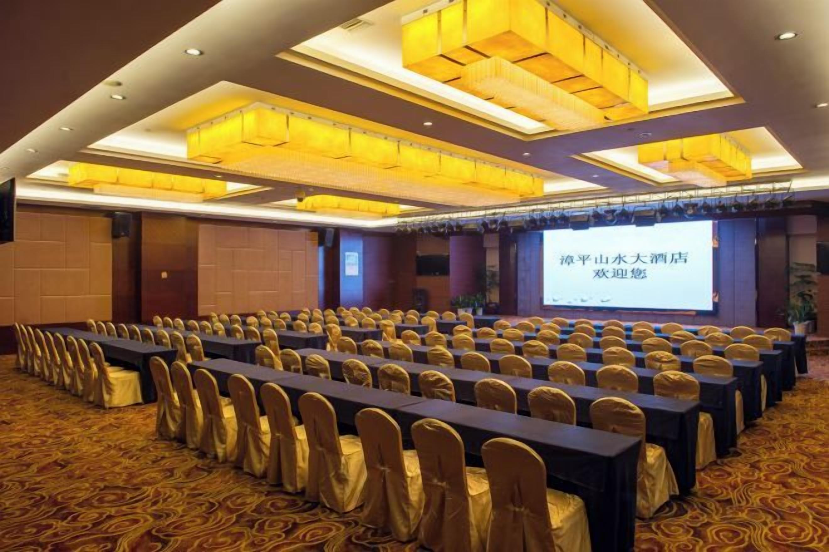 Un salón de conferencias con filas de mesas y sillas, una pantalla grande y un candelabro.