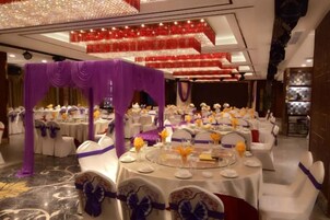Un salón de banquetes con mesas redondas, sillas blancas y cortinas moradas.