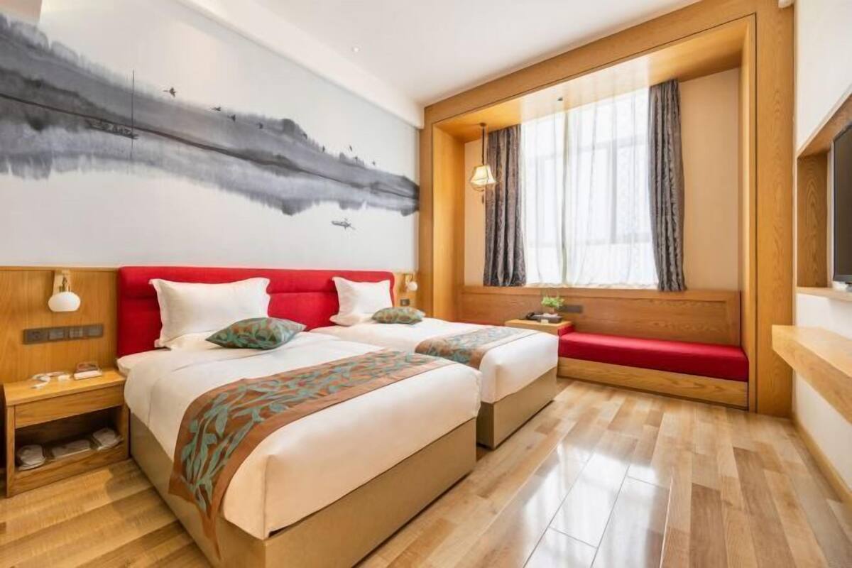 Ein Hotelzimmer mit einem großen Bett, einem roten Kopfteil, einer hölzernen Bank, einem Wandbild und einem Fenster mit Vorhängen.