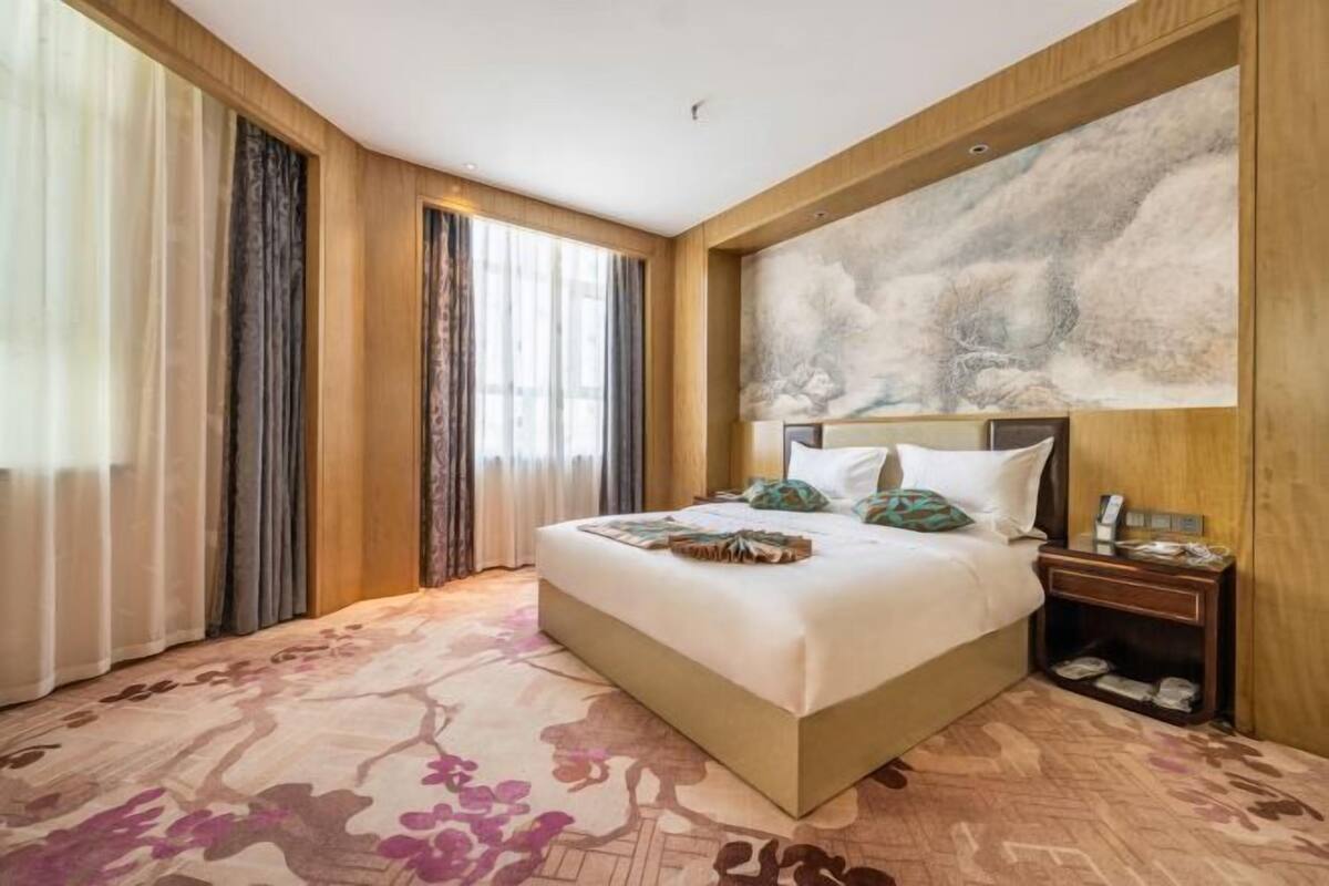 Ein Hotelzimmer mit einem großen Bett, einem an der Wand befestigten Kopfteil mit einem Gemälde, einem Nachttisch und einem Teppich mit floralem Muster.