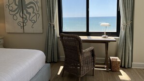 A bedroom with a bed, a desk with a lamp, a wicker chair, and a view of the sea through a large window. - SOPHISTICATED BEACHFRONT RETREAT -  New Furniture & Decor!! (Orange Beach)