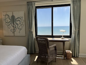 Un dormitorio con cama, escritorio con lámpara, silla de mimbre y vista al mar a través de un ventanal grande.