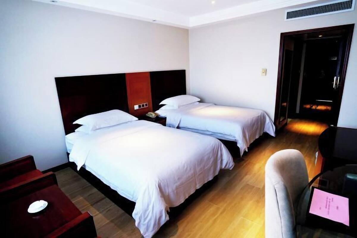 A hotel room with two single beds, a wooden floor, a red side table, and a desk with a chair.