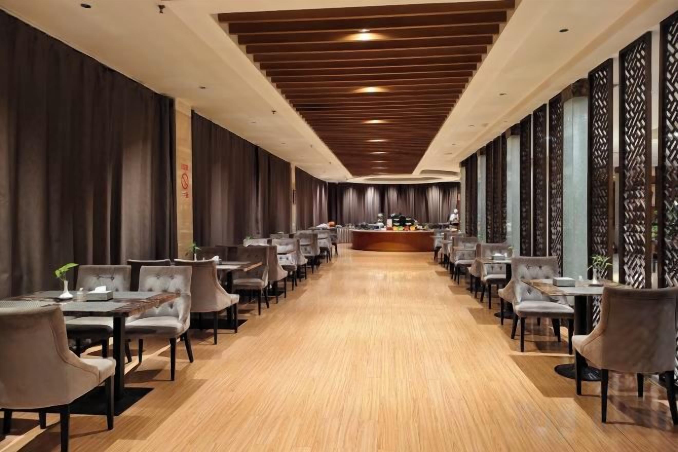 A long, narrow hotel lobby with a central reception desk, rows of tables and chairs, and a wooden floor.