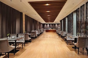 Un vestíbulo de hotel largo y estrecho con una recepción central, filas de mesas y sillas, y piso de madera.