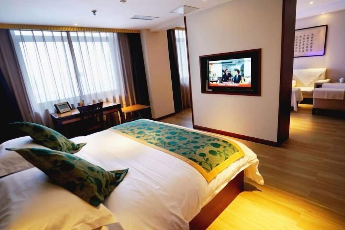 A hotel room with a large bed, a TV on the wall, a desk with a chair, and a window with curtains.