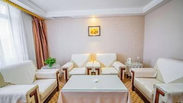 A hotel room with a checkered floor, a central glass table, two white sofas, a small round table with a lamp, and a framed picture on the wall.