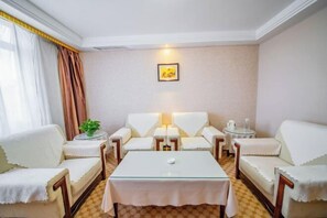 A hotel room with a checkered floor, a central glass table, two white sofas, a small round table with a lamp, and a framed picture on the wall.