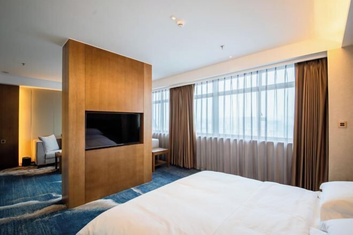 A modern hotel room with a large bed, a flat-screen TV mounted on a wooden panel, a seating area with a sofa and armchair, and a large window with curtains.