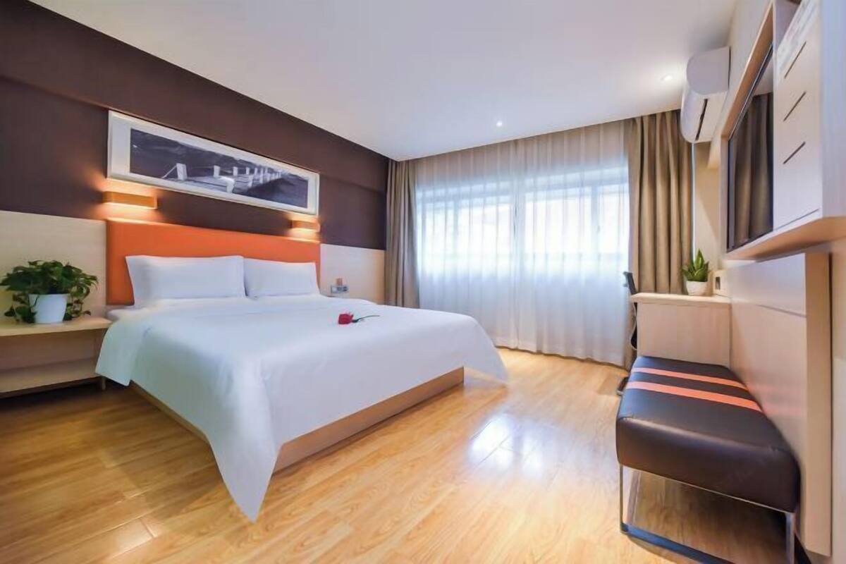 Moderni hotellihuone, jossa on suuri sänky, yöpöytä, jossa on kasvi, työpöytä tuolilla ja ikkuna, jossa on verhot.