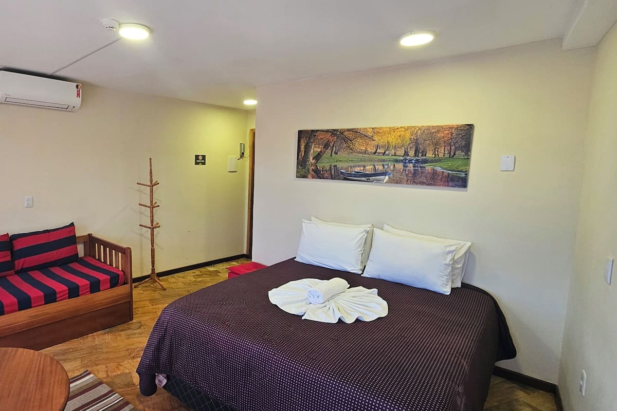 Una habitación con cama, sofá, una silla de madera, una mesita, aire acondicionado y un cuadro en la pared.
