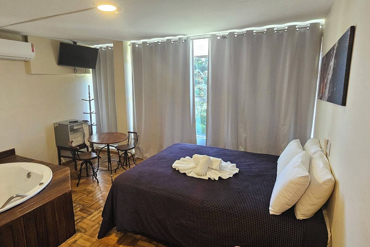 Habitación de hotel con una bañera grande, una cama con una toalla doblada cuidadosamente, una mesita con sillas y un televisor montado en la pared.