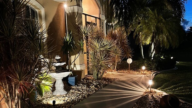 Een goed verlicht pad dat naar een gebouw leidt, met een waterpartij, omgeven door palmbomen en een aangelegde tuin.
