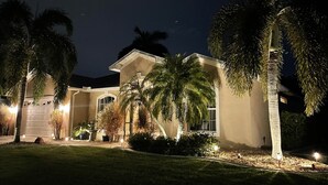 Een goed verlicht woonhuis met garage, palmbomen en een aangelegde tuin, 's nachts.