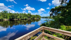 A wooden deck overlooking a calm river with dense greenery on both sides. - Peaceful property on the historic Suwannee River. (Bell)