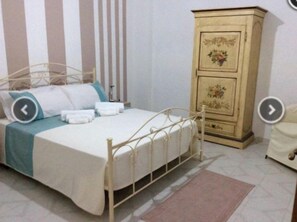 Una cama bien hecha con estructura de metal, un aparador de madera con detalles florales y una pared a rayas al fondo.